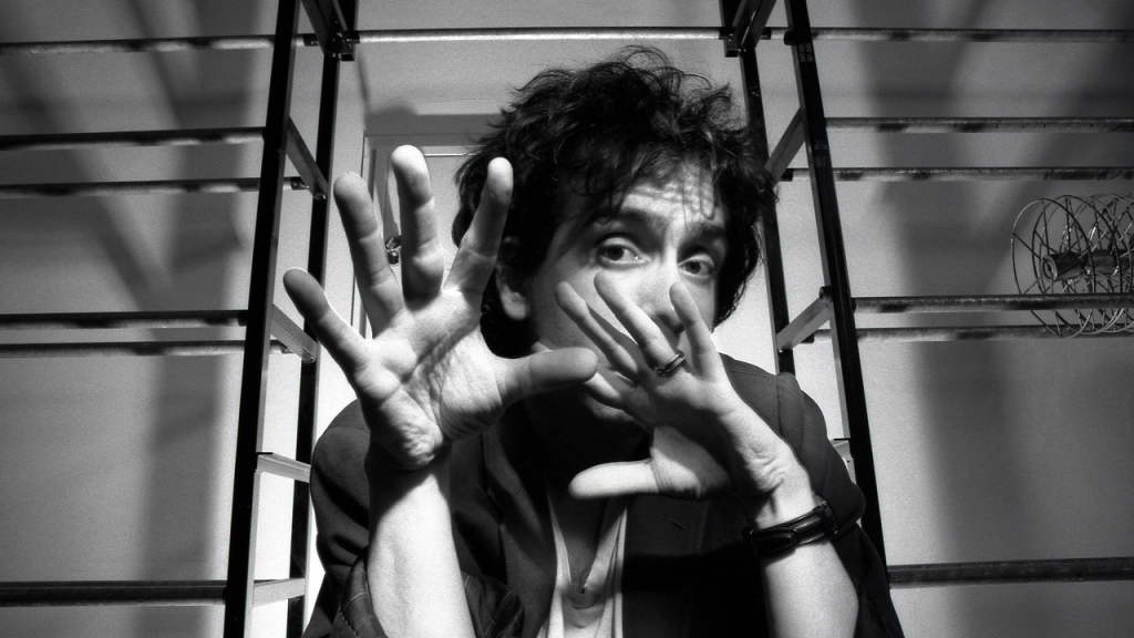 Person with tousled hair holds hands up in front of face, fingers spread wide, sitting between metal frames in a black and white photo.