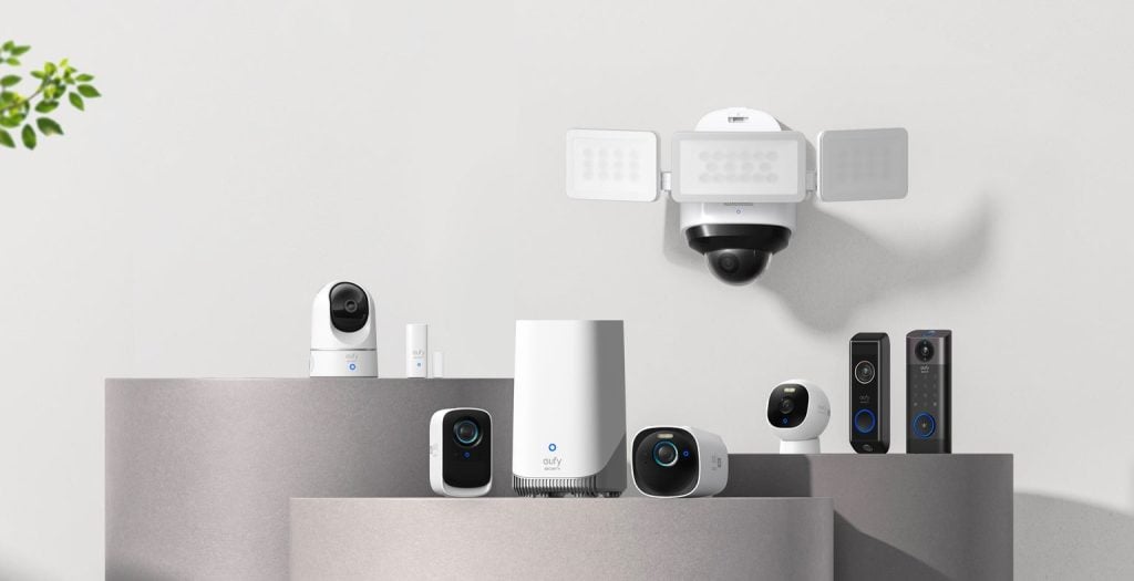 A display of various home security devices, including cameras, a floodlight camera, video doorbells, and a central hub, arranged on gray platforms against a plain wall.