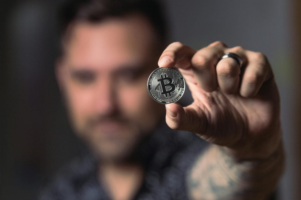 A person holds a physical Bitcoin token up close to the camera, with their face blurred in the background.