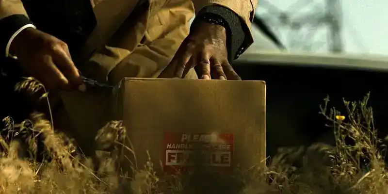 A person in a suit crouches outdoors, using a knife to open a cardboard box labeled "FRAGILE" with dry grass in the foreground.