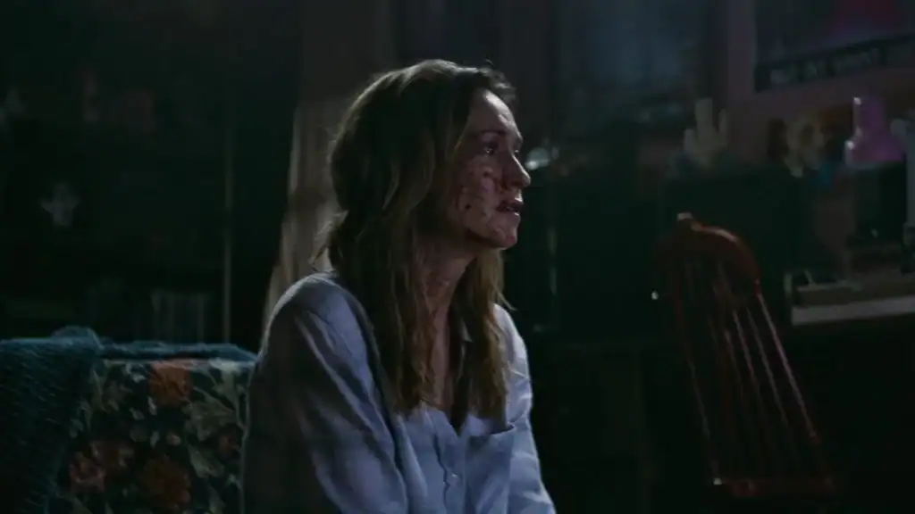A woman with long hair and blood on her face sits on a couch in a dimly lit room, looking to the side.