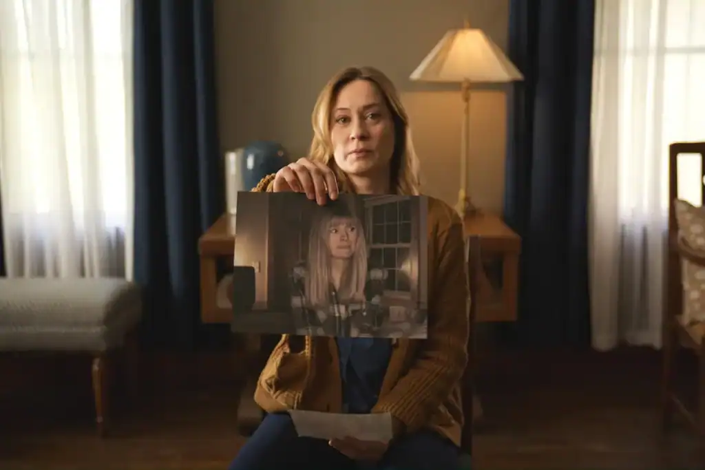 A woman sits indoors, holding up a printed photo of another woman. A lamp, desk, and curtains are visible in the background.