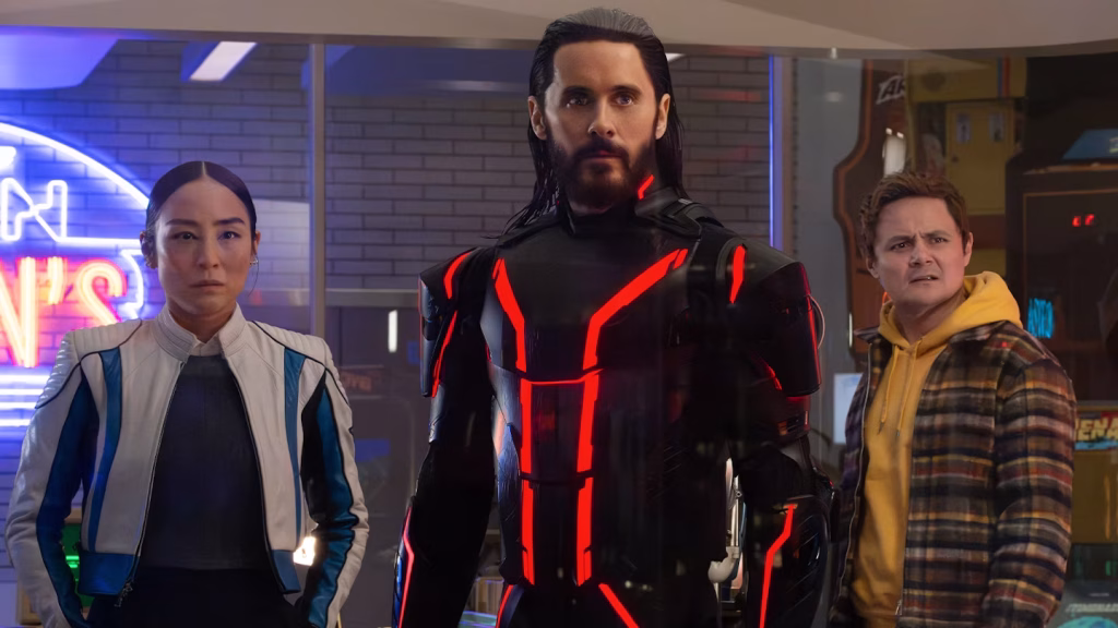 Three people stand indoors: a serious woman in a white and blue jacket, a man in a black suit with red neon lines, and another man in a yellow hoodie and plaid jacket looking confused.