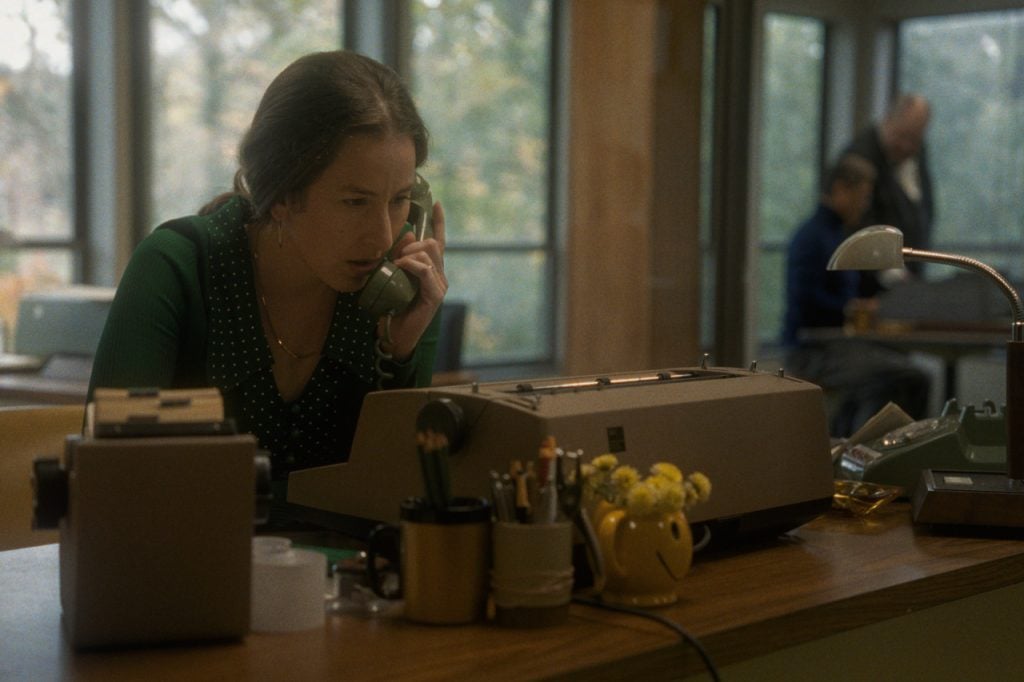 A woman sits at a desk working on a typewriter while talking on a corded phone; office supplies and a yellow smiley-face mug are on the desk, with two men in the background.