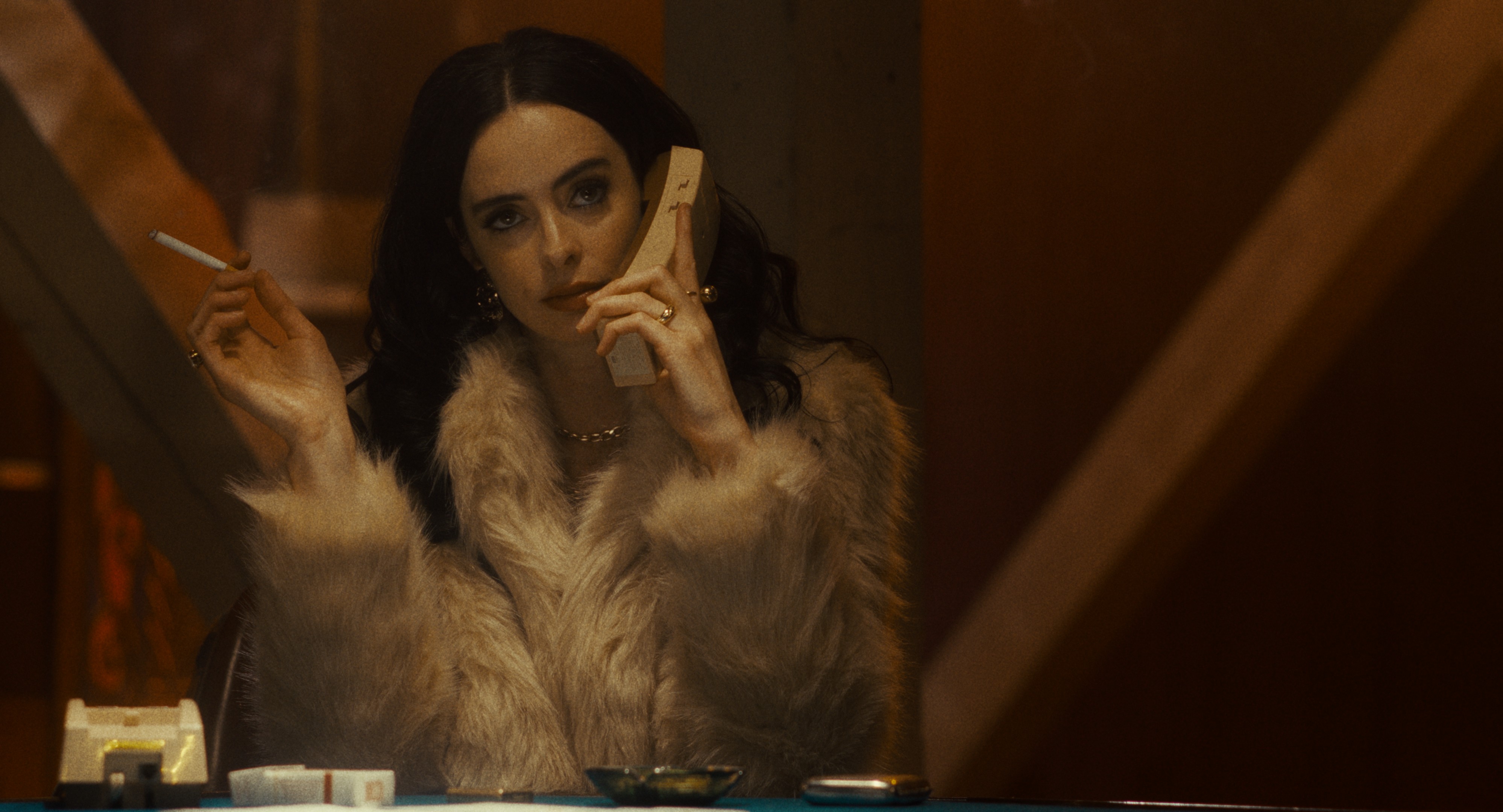 A woman in a fur coat sits at a table, holding a cigarette in one hand and a beige telephone receiver in the other, looking off to the side.