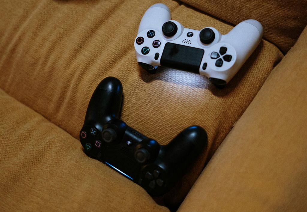 A white and a black video game controller are placed on a light brown fabric sofa.