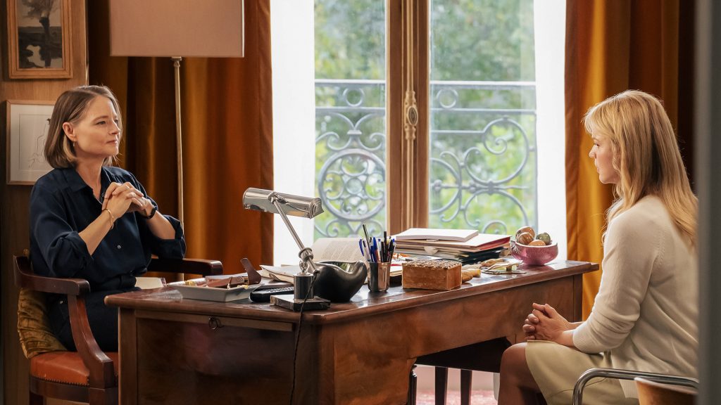 Two women sit across from each other in an office, separated by a wooden desk with papers, books, and a lamp. One woman listens attentively while the other speaks.