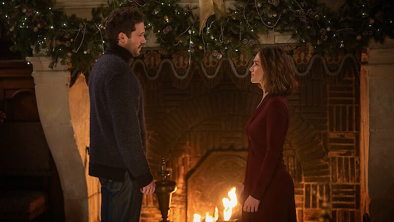A man and a woman stand facing each other in front of a lit fireplace, with holiday garland and lights decorating the mantel above them.