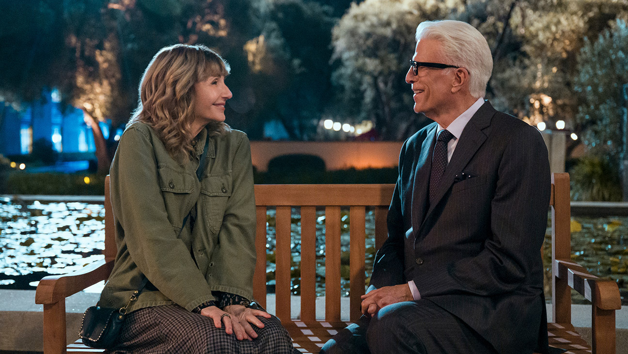 A woman in casual clothes and a man in a suit sit on a wooden bench by a pond at night, smiling and talking.
