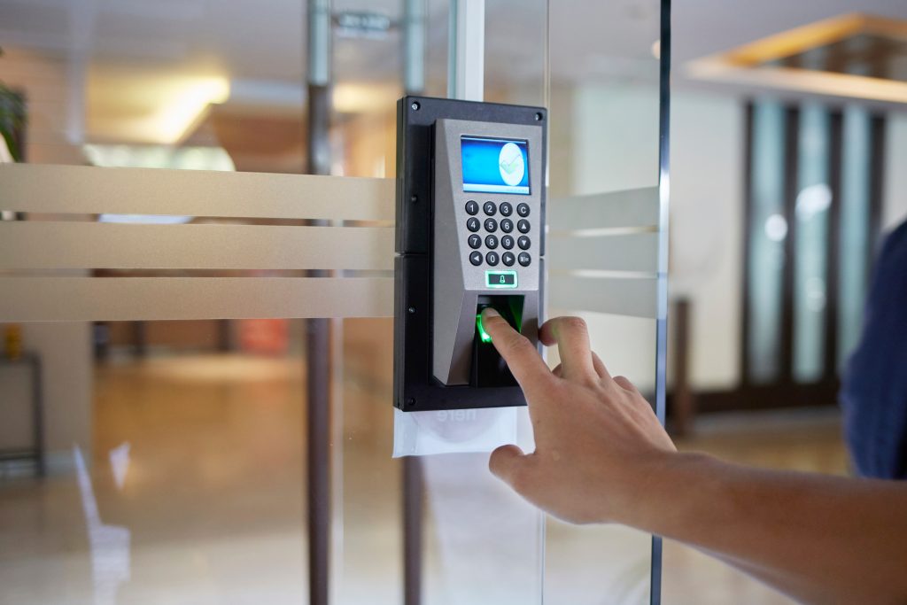A person places their finger on a biometric fingerprint scanner attached to a glass door for access control in a modern building.