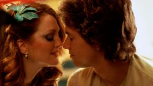 A woman with a blue flower in her hair and a man sit close together, nearly touching noses, in a softly lit setting.