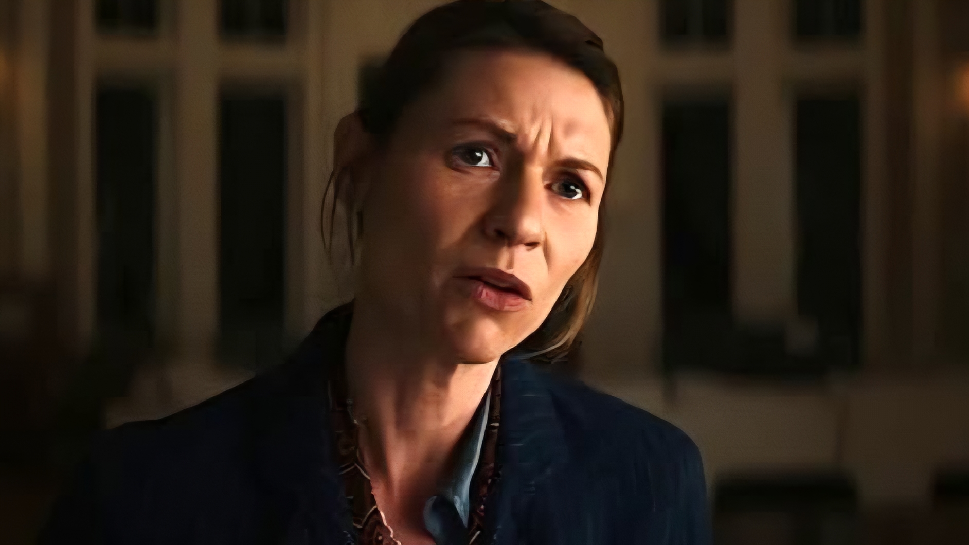 A woman with light brown hair pulled back looks concerned while speaking indoors, dressed in a dark blazer and patterned blouse.