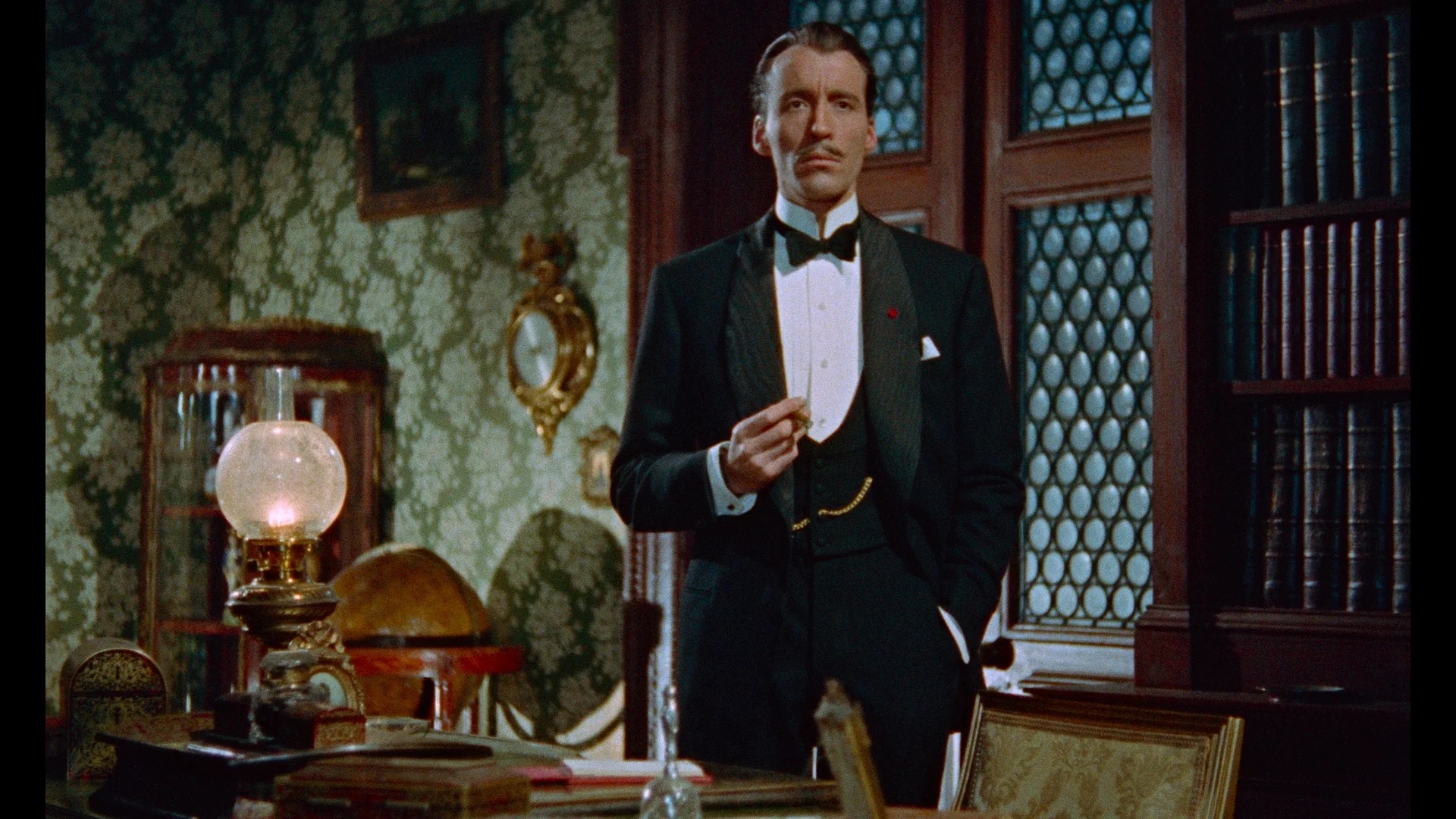 A man in a tuxedo stands in an ornate, antique-furnished study, holding a small object in one hand, with bookshelves and patterned wallpaper in the background.