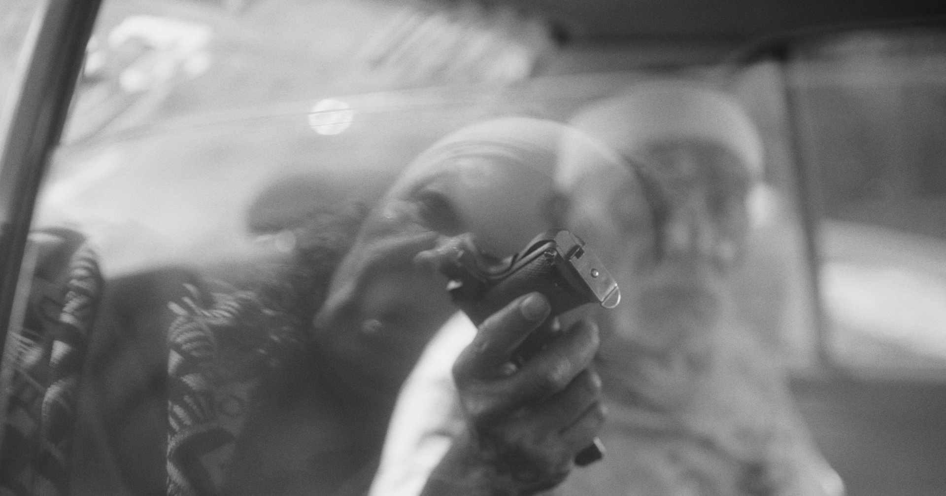 A person holds a handheld device near their face while sitting in a car, viewed through a window with reflections.