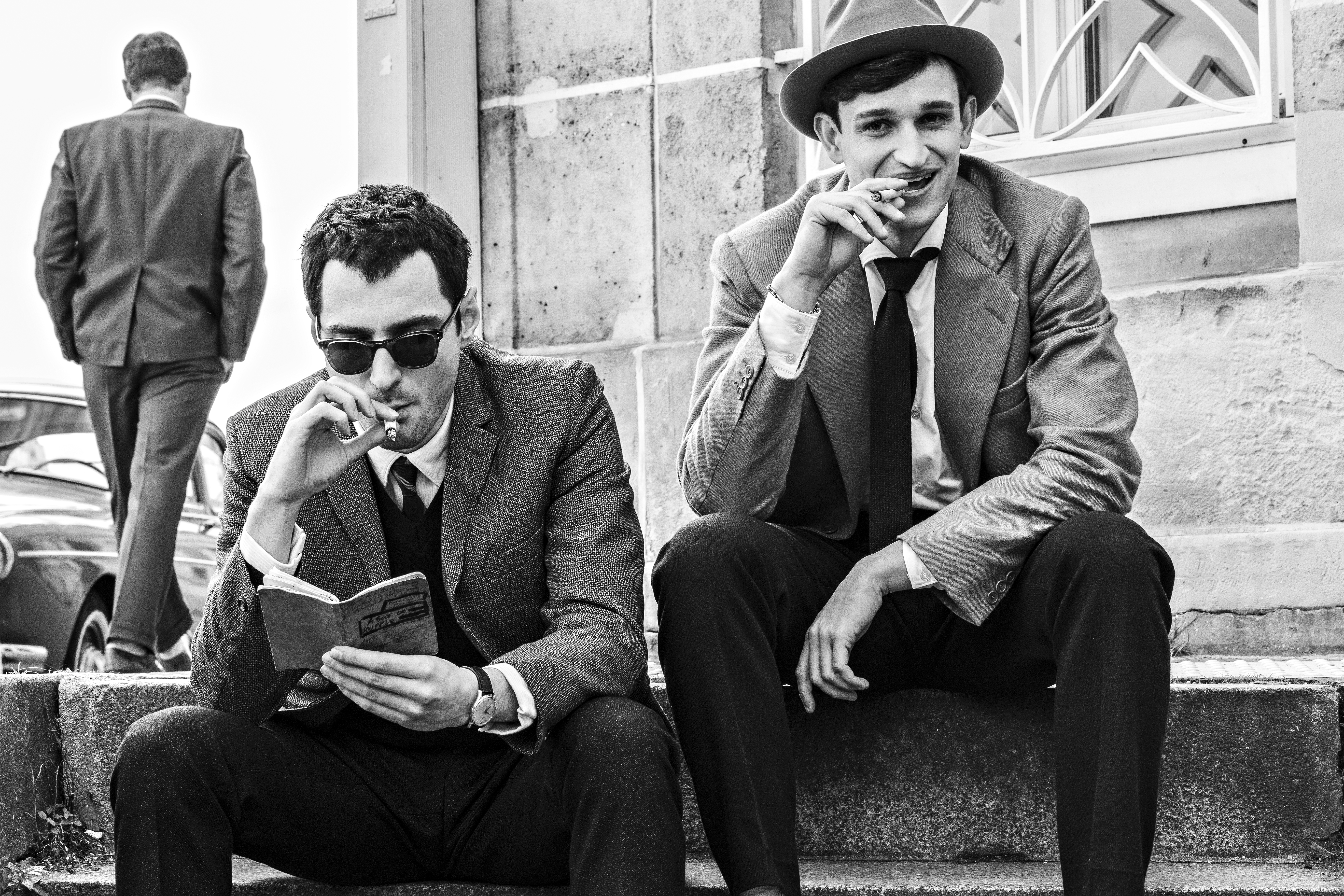 Two men in suits sit on steps; one reads a book while smoking, the other smiles and smokes. A third man, also in a suit, walks away in the background. The image is black and white.