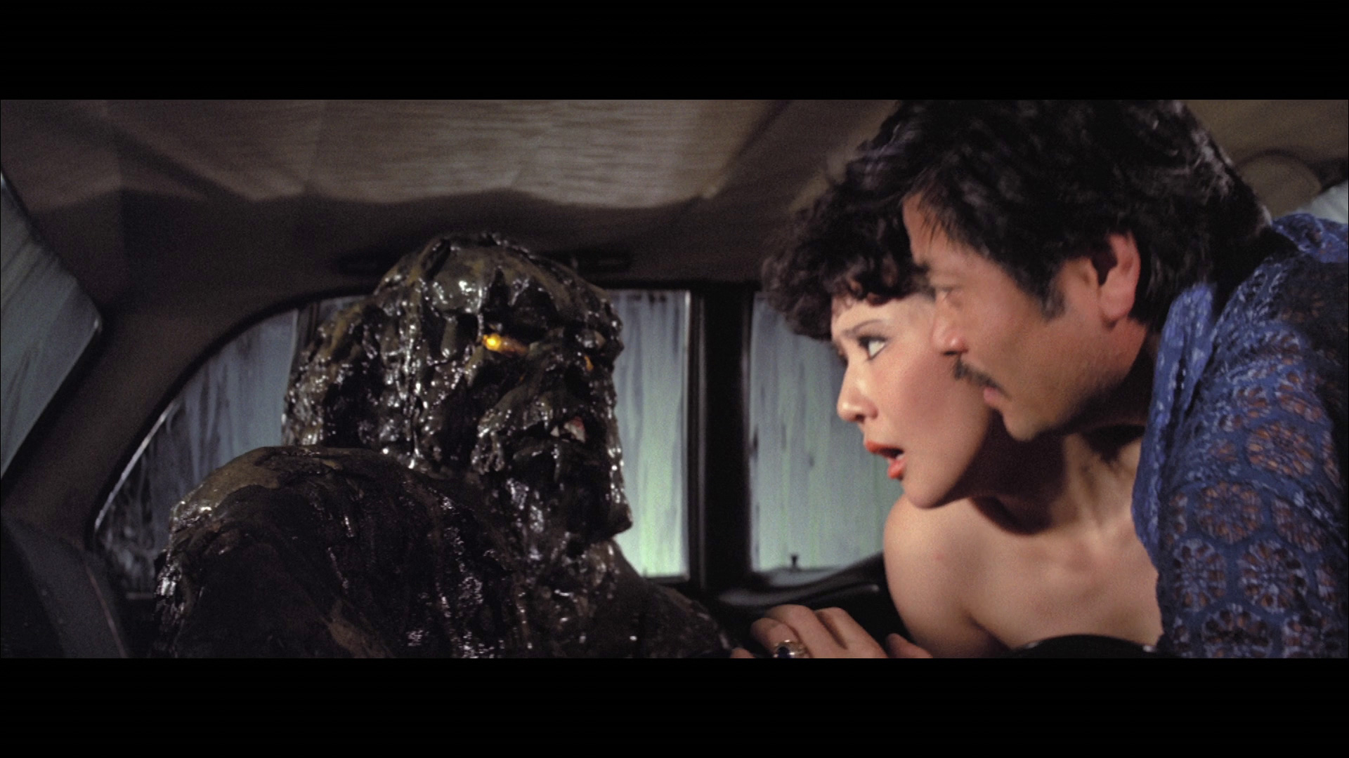 Two people in a car react with concern as a dark, mud-covered creature with glowing eyes sits beside them.