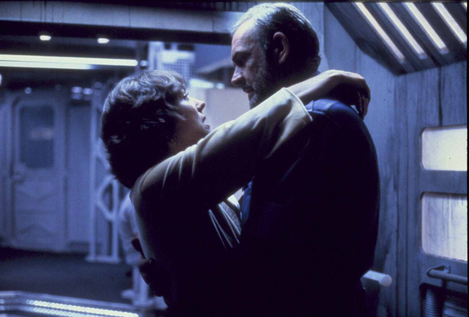 Two people embrace closely in a dimly lit, industrial-looking room with metallic walls and overhead lights.