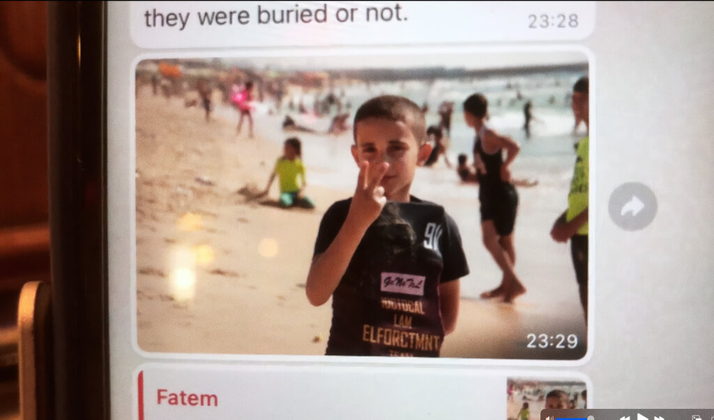A boy stands on a crowded beach making a peace sign with his fingers, while other people are visible in the background near the water.