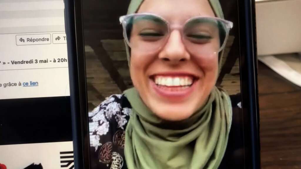 A person wearing glasses and a green hijab smiles broadly during a video call, with a computer screen visible in the background.