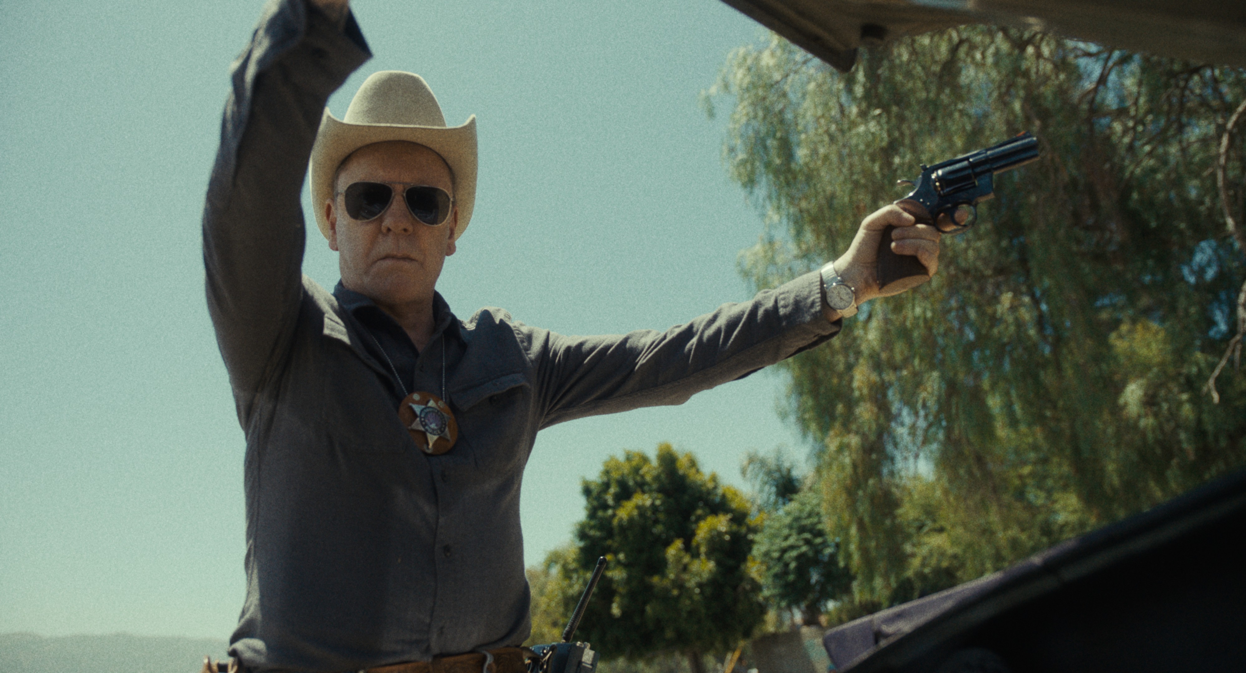 A sheriff in a cowboy hat and sunglasses points a revolver while raising his other hand, outdoors with trees in the background.