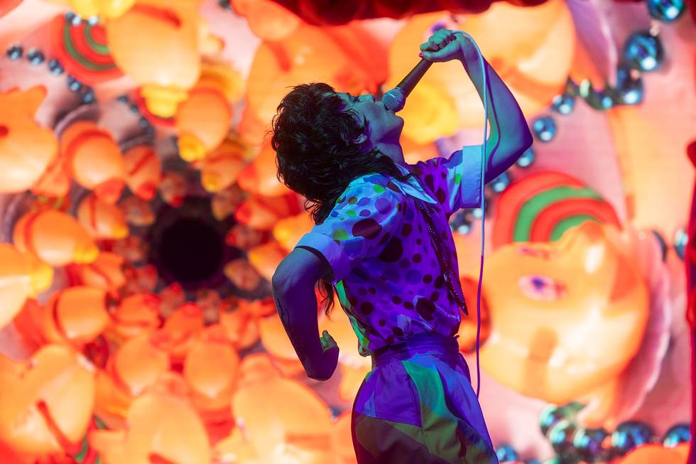 A performer sings into a microphone in front of a vibrant, kaleidoscopic background featuring colorful abstract shapes.