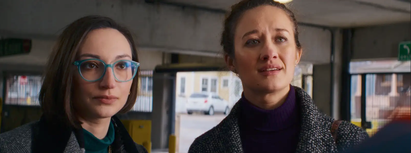 Two women in coats stand indoors, one with blue glasses and short hair, the other with her hair tied back, both looking slightly upward.