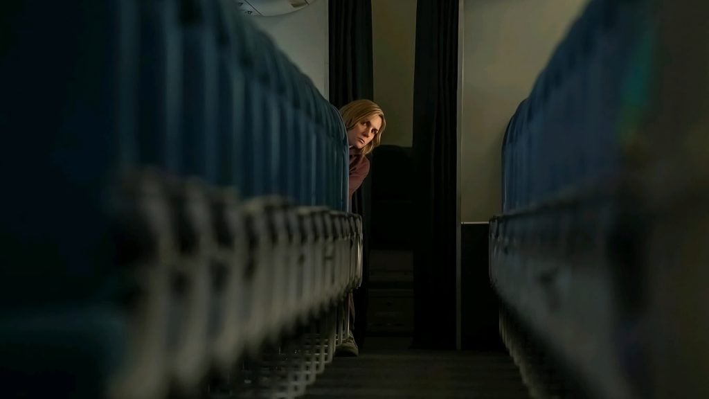 A woman stands in the aisle between rows of empty airplane seats, looking out from behind a curtain.
