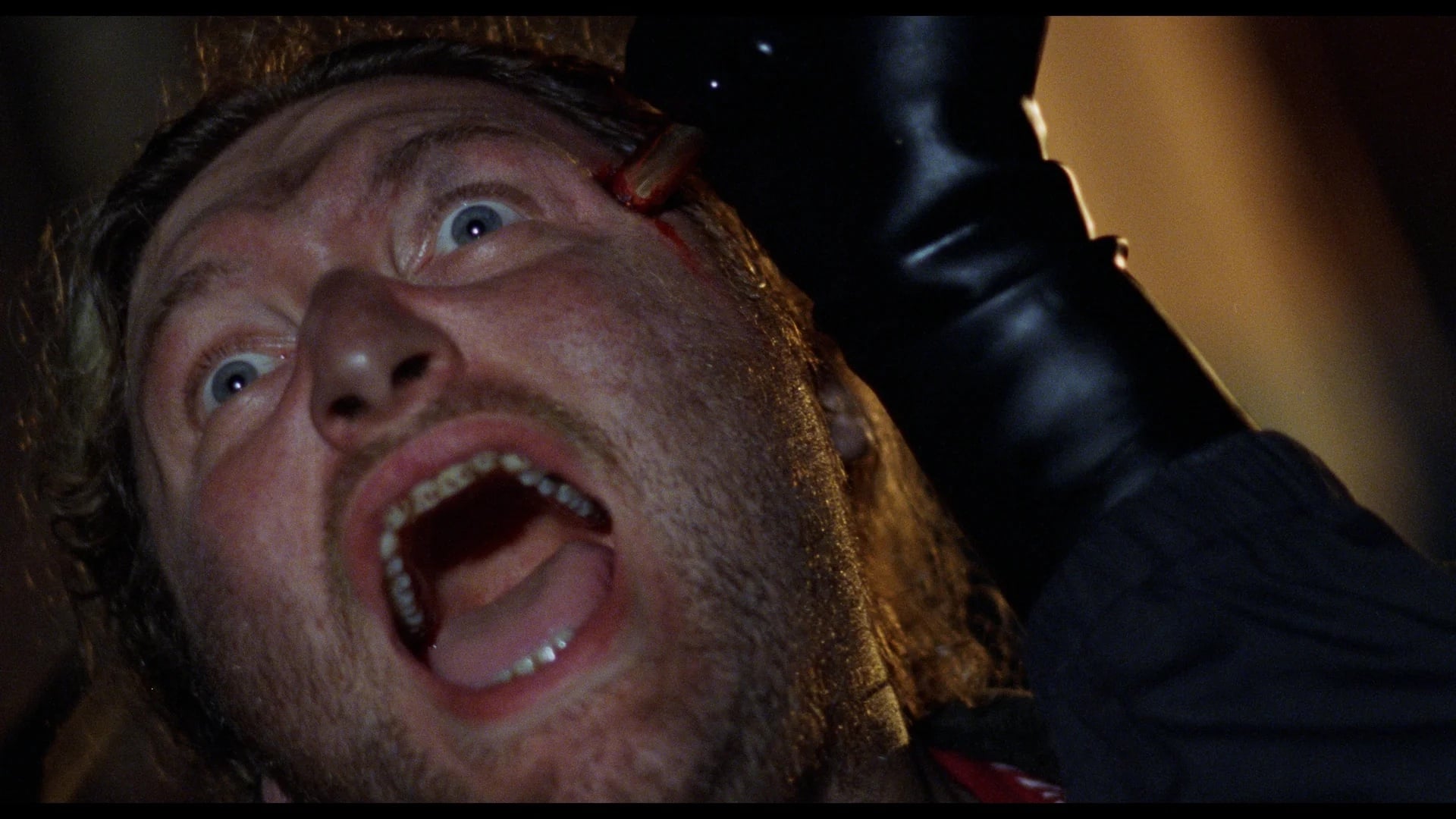 A close-up of a man screaming in fear as a black-gloved hand presses a gun to his head.
