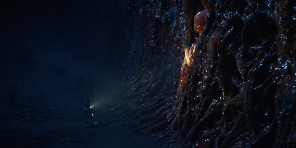 A person holding a flashlight stands near a large, slimy, organic wall with glowing, human-like figures embedded in its surface in a dark, eerie environment.