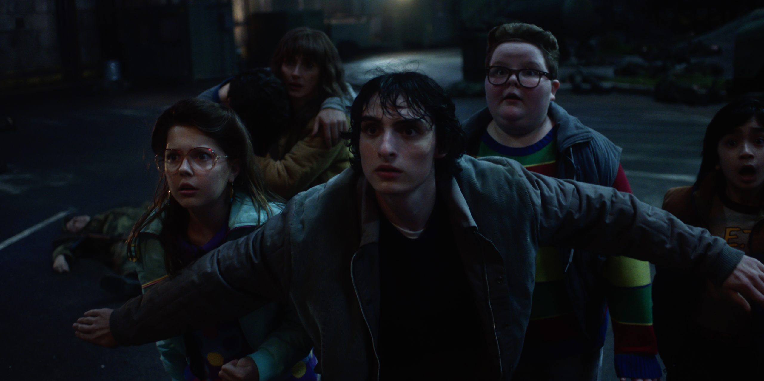 A group of six teenagers stand close together outdoors at night, looking ahead with tense and concerned expressions. One boy extends his arms protectively in front of the group.