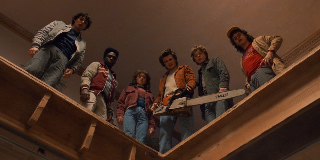Six teenagers stand on a wooden landing looking down, one holding a chainsaw, with a beige ceiling visible above them.