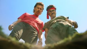 Two men with mustaches, one in a red shirt and the other in green with a red headband, look down into a hole against a blue sky background.