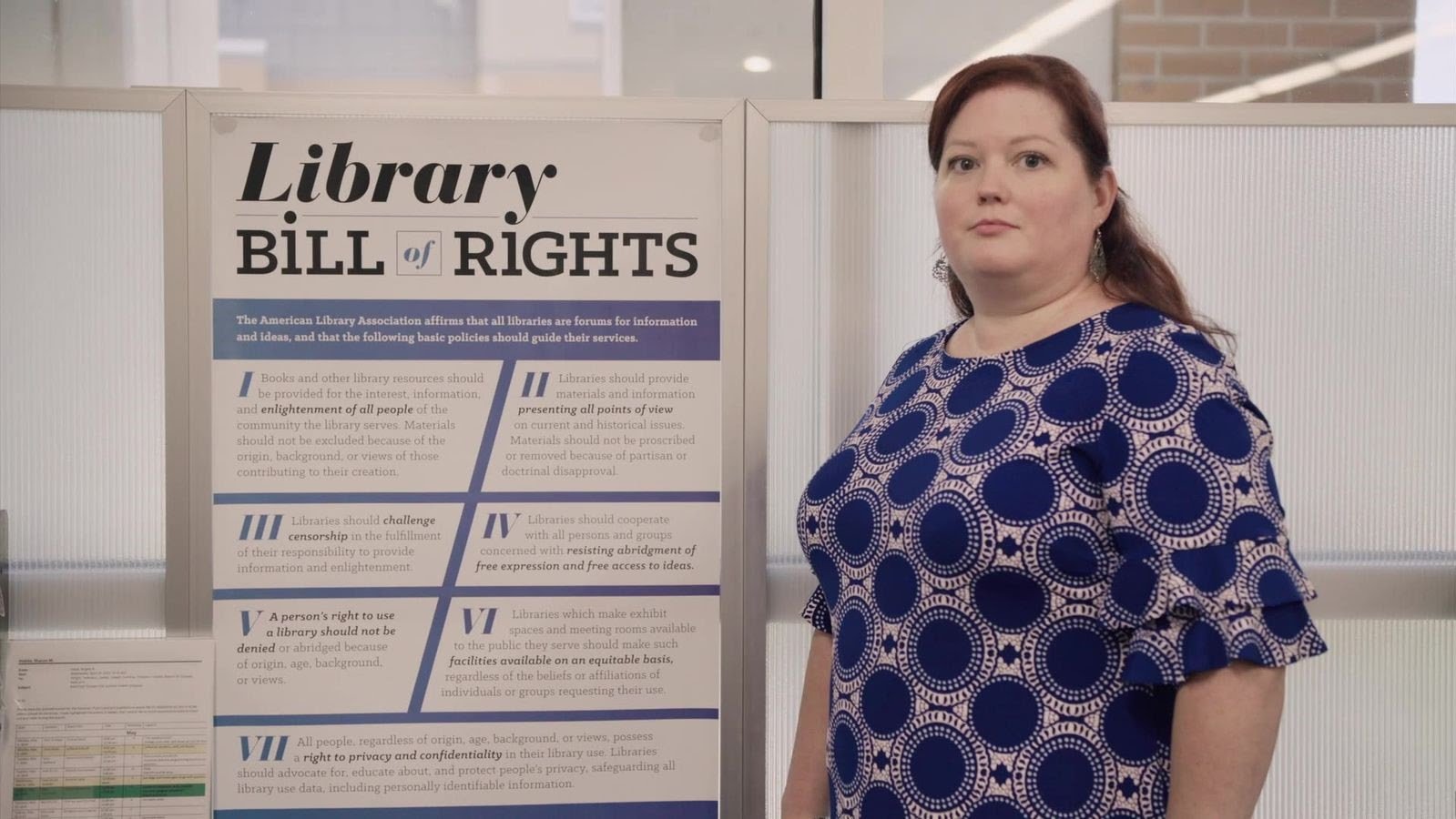 A woman in a blue patterned dress stands next to a poster titled "Library Bill of Rights" in an indoor setting.