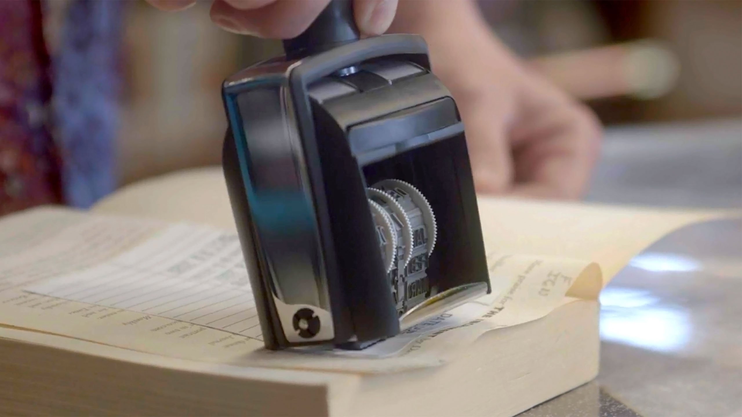 A hand uses a date stamp to mark a slip of paper inside a book, likely for library record-keeping or due date tracking.