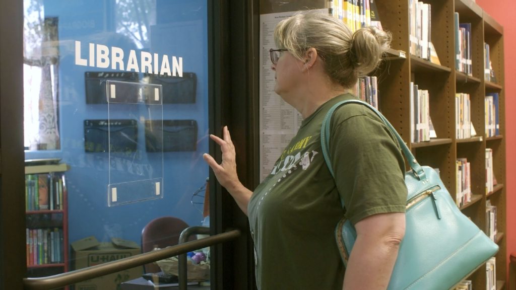 A woman with a turquoise bag stands at a glass door labeled "Librarian," looking inside a library office.