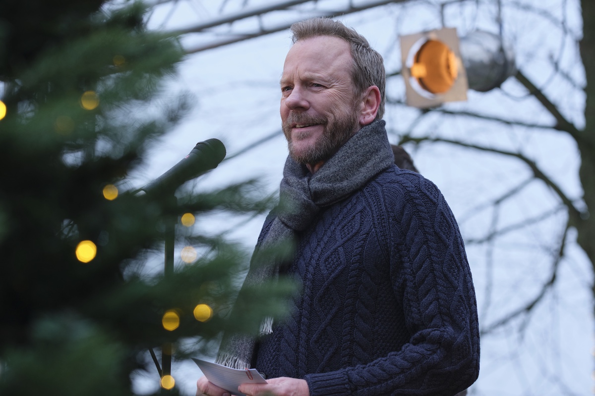 A man in a dark sweater and scarf speaks into a microphone outdoors, near a decorated tree with string lights.