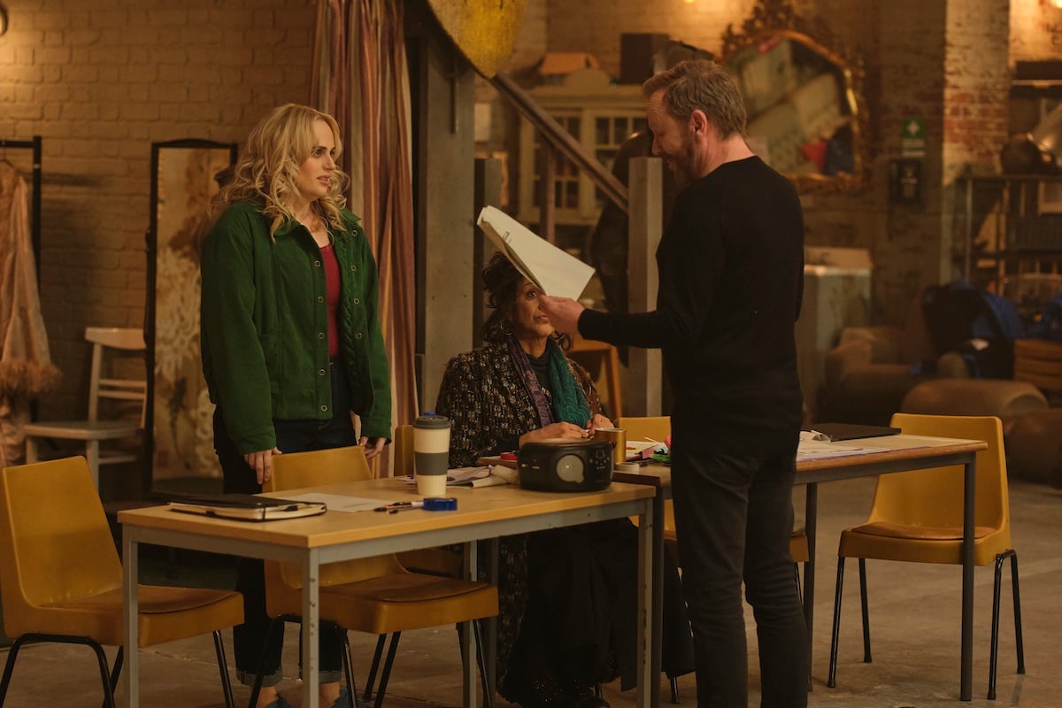 Three people are gathered around a table in a rustic room; one man stands holding papers while two women sit and listen. The table is covered with office supplies and a speaker.