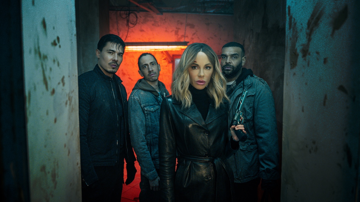 Four people stand in a dimly lit, grungy hallway with red lighting; the woman in front wears a black coat, while the three men behind her wear dark clothing.
