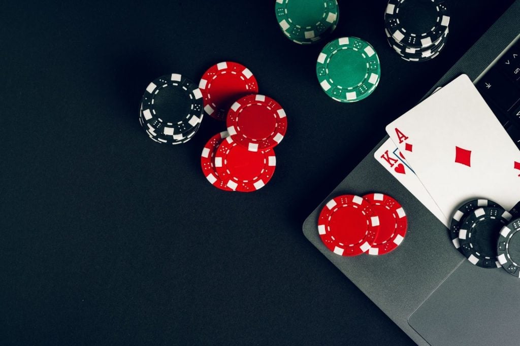 Poker chips and playing cards, including ace of spades and ace of diamonds, are placed on a laptop keyboard against a dark background.