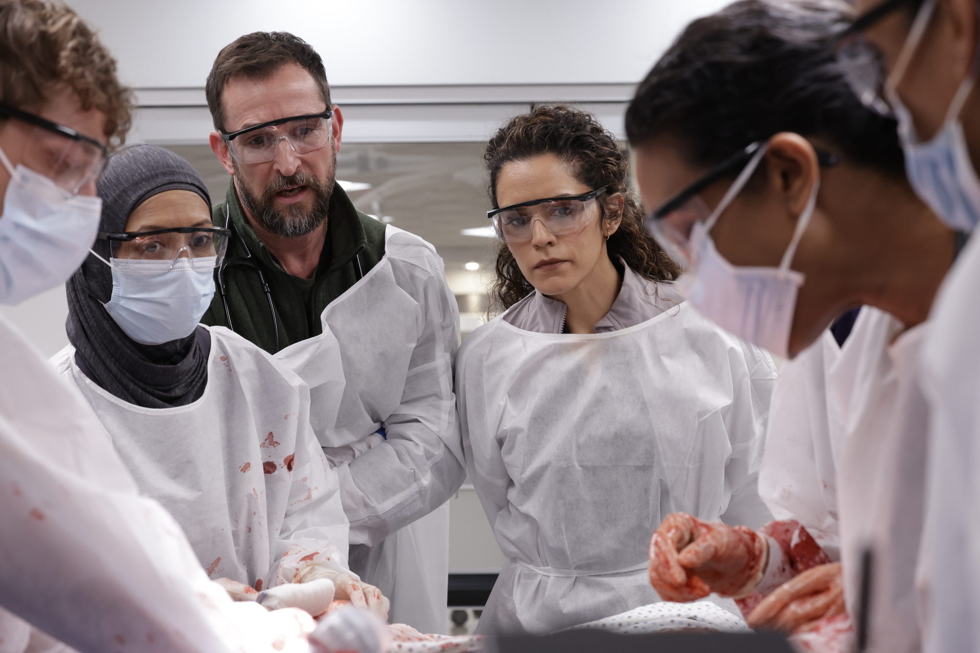 A group of people in protective gowns, masks, and goggles observe and assist with a surgical procedure in a medical setting.