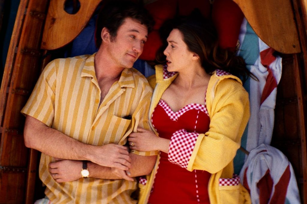 A man and woman lie side by side in a wooden boat, gazing at each other. The man wears a yellow striped shirt; the woman wears a red dress with a yellow robe.