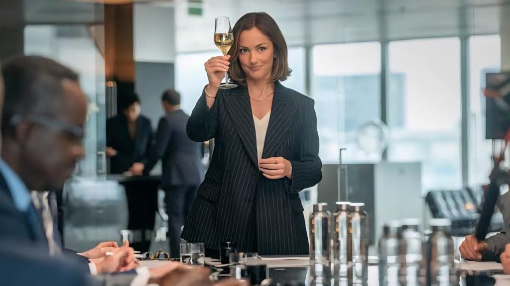 A woman in a business suit stands holding up a glass of white wine in an office meeting room, with seated colleagues in the foreground.