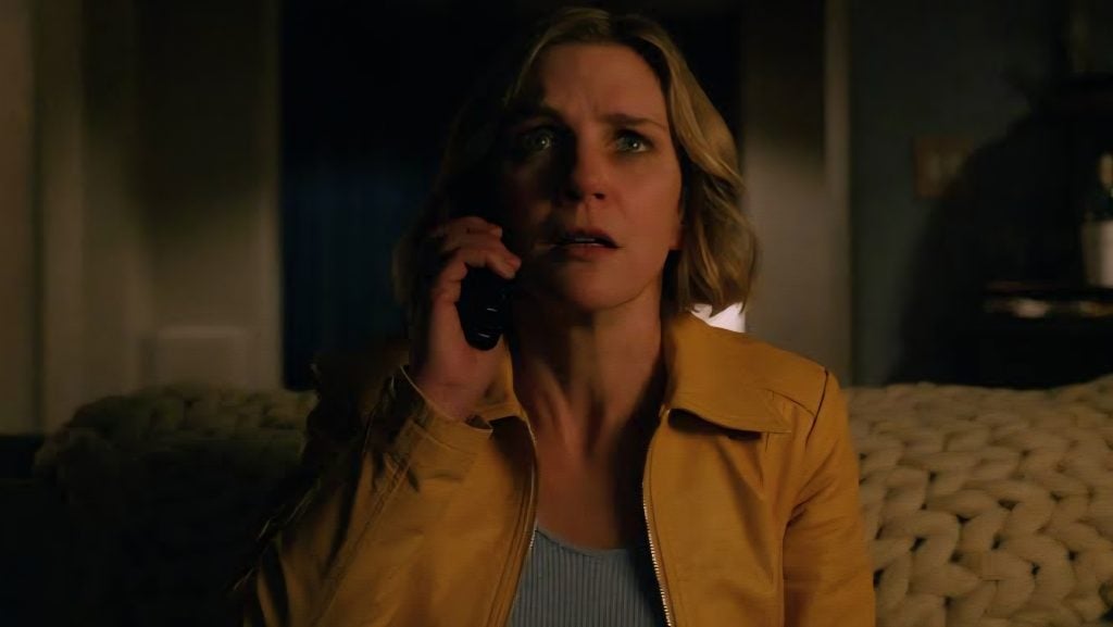 A woman with blonde hair in a yellow jacket sits on a couch, looking concerned while holding a phone to her ear in a dimly lit room.