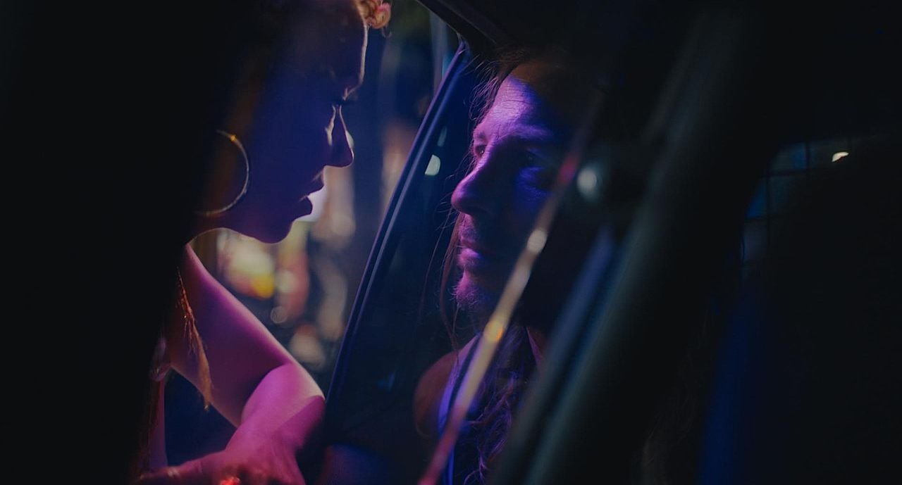 Two people have an intense conversation through a car window at night, illuminated by colorful lights.