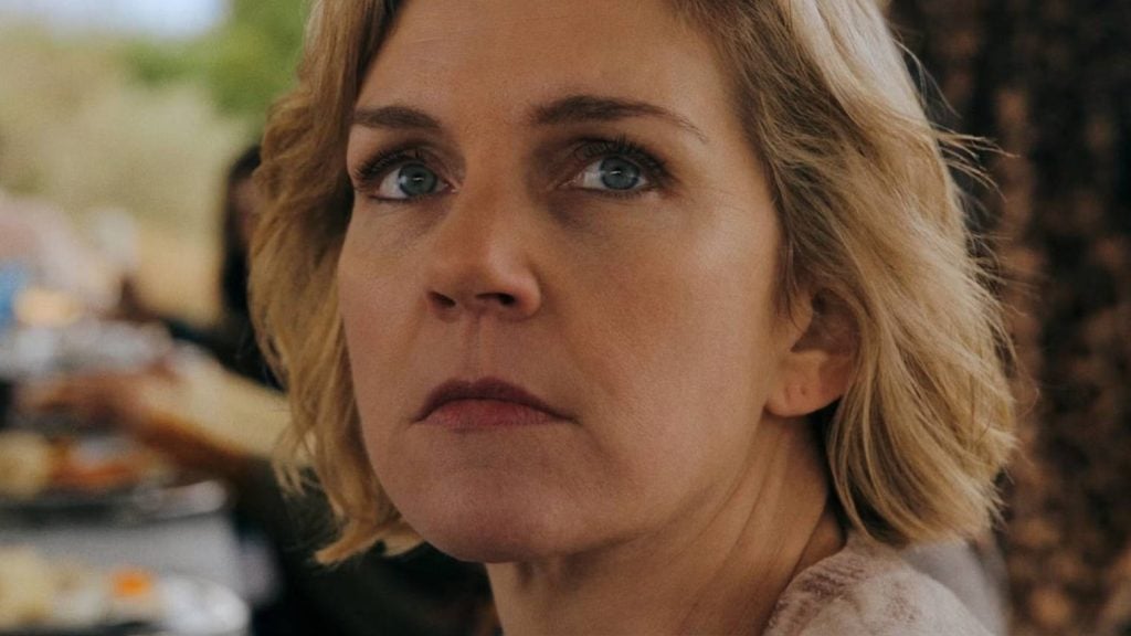 A blonde woman with short hair and blue eyes looks slightly upward with a serious expression; background is softly blurred.