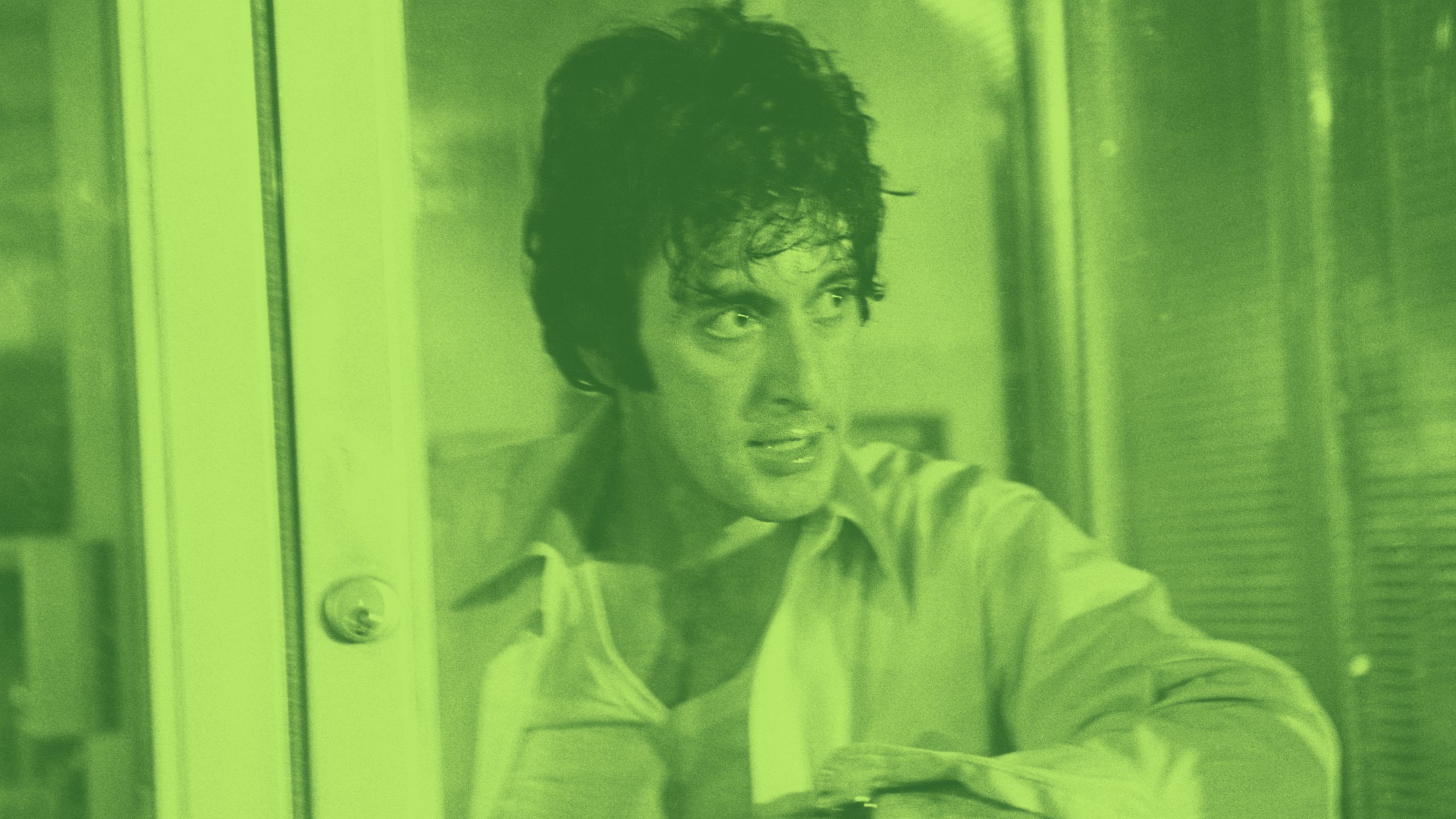 A man with curly dark hair and an intense expression stands by a glass door. The image has a green filter.