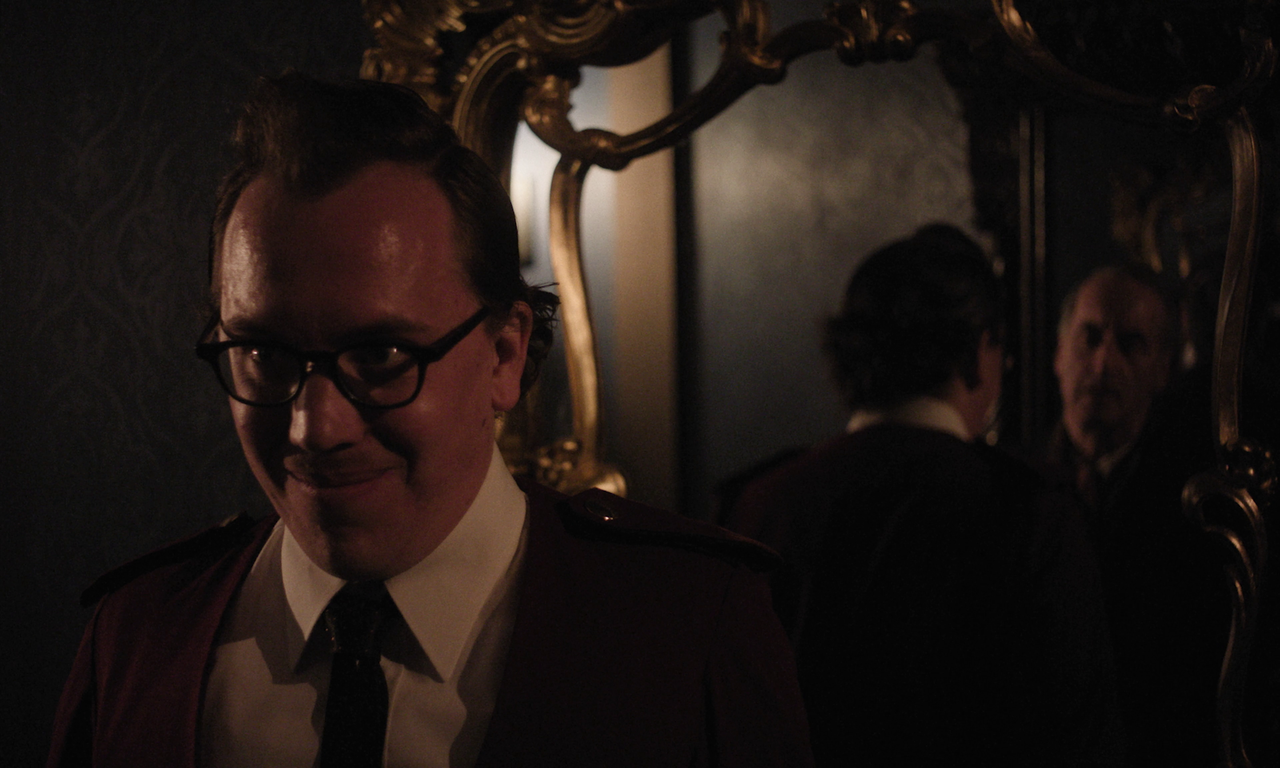 A man in glasses and a suit with a dark tie stands in a dim room, smirking, with his reflection seen in an ornate gold-framed mirror behind him.
