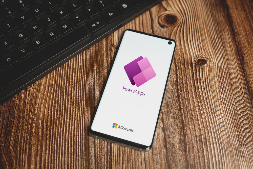 A smartphone displaying the Microsoft PowerApps logo on its screen rests on a wooden surface next to a black keyboard.