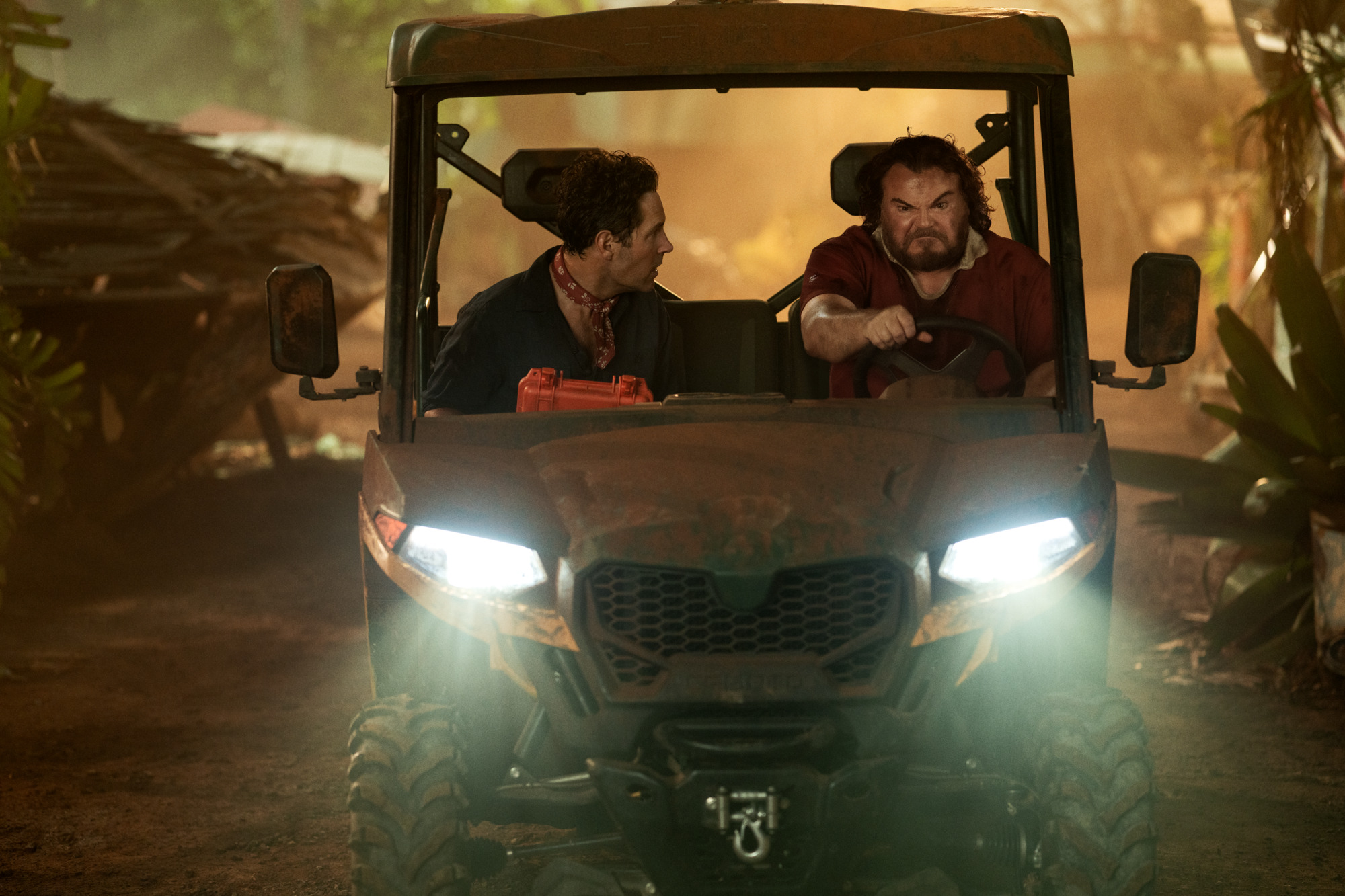 Two men are sitting in the front seats of an off-road utility vehicle in a jungle setting, with one man driving and the other looking at him.