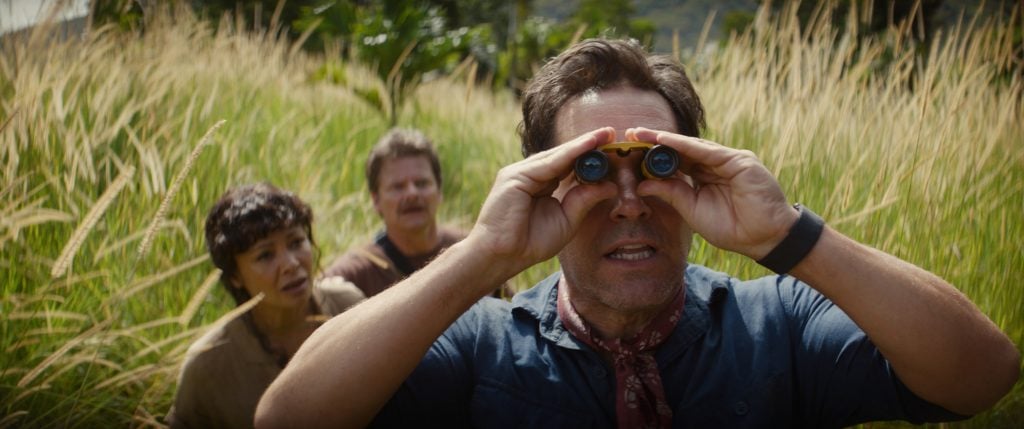 Three people stand in tall grass; the person in front is looking through binoculars, while the two behind watch attentively.